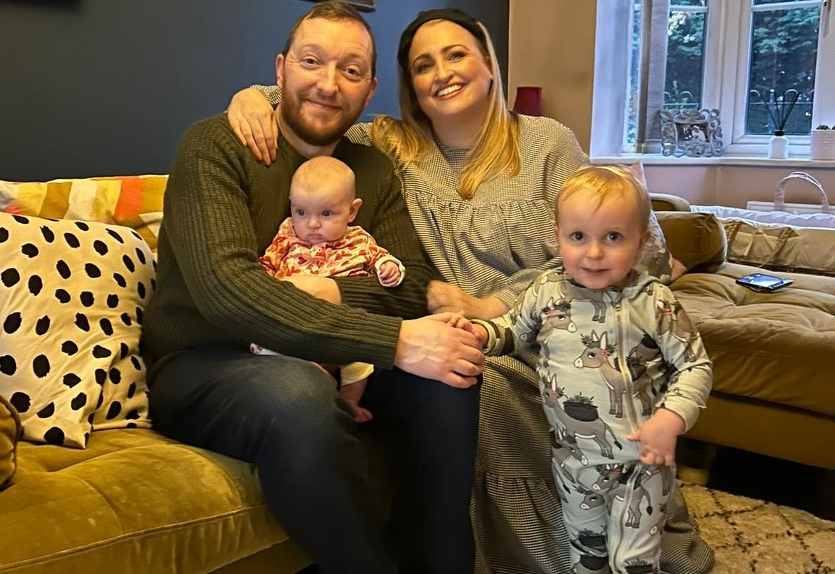 Image shows Hannah and Darren on the sofa of their home with a baby on their lap and toddler standing, all (except baby!) smiling for the camera. 