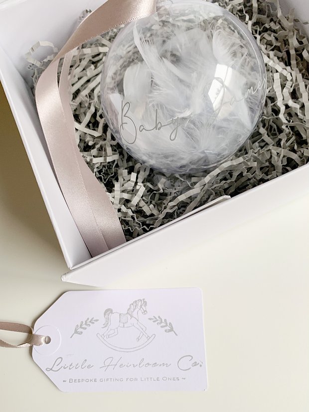 A clear bauble filled with white feathers and personalised with silver italic writing