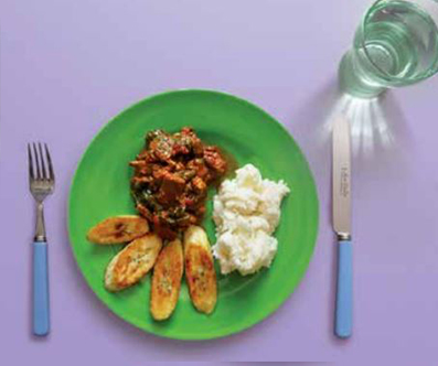 Image of stew on a plate with plantains and mashed potato. There is glass of water and cutlery on the table. 