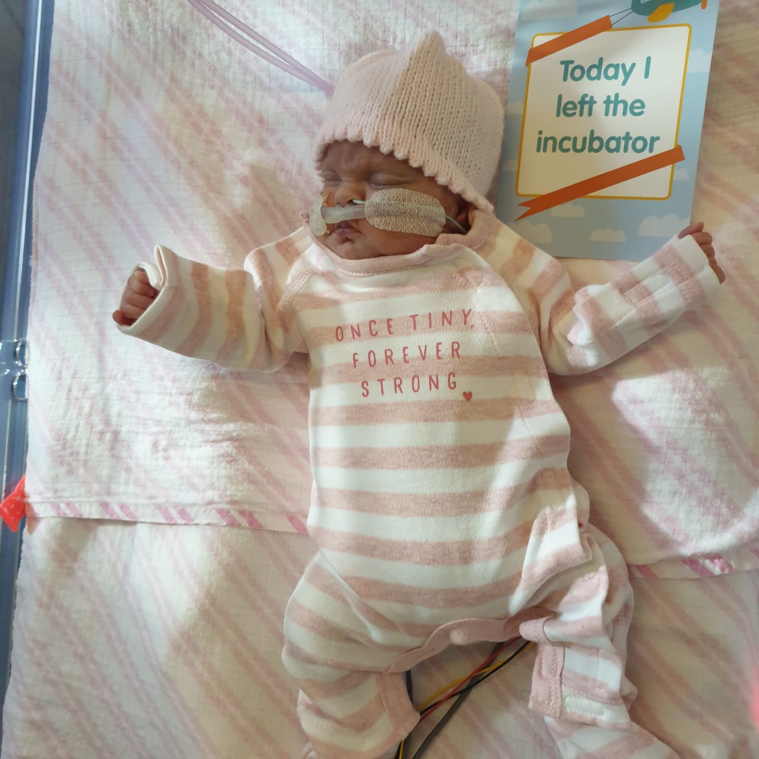 A baby girl lies on her back in a sleep suit reading 'Once tiny, forever strong', next to a sign reading 'Today I left the incubator'