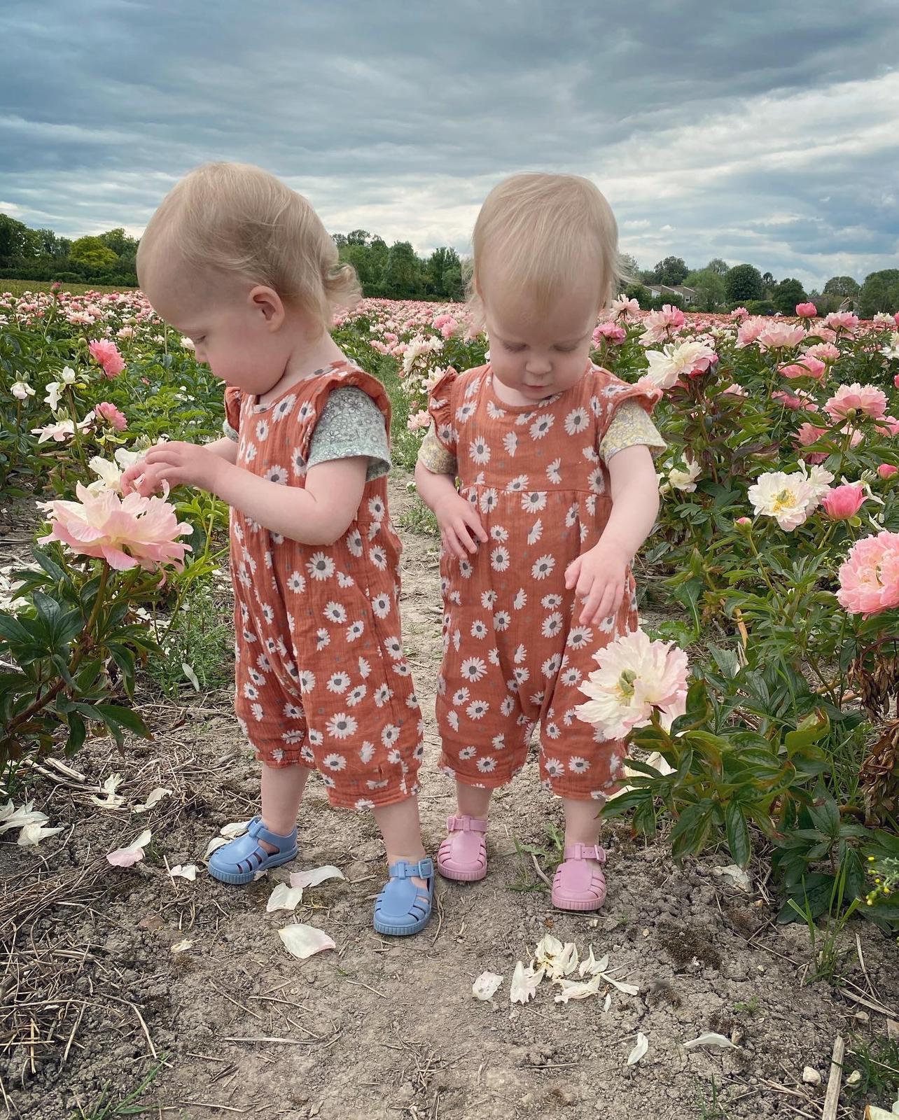 Twin girls wearing matching floral romper suits, standing in a field of flowers