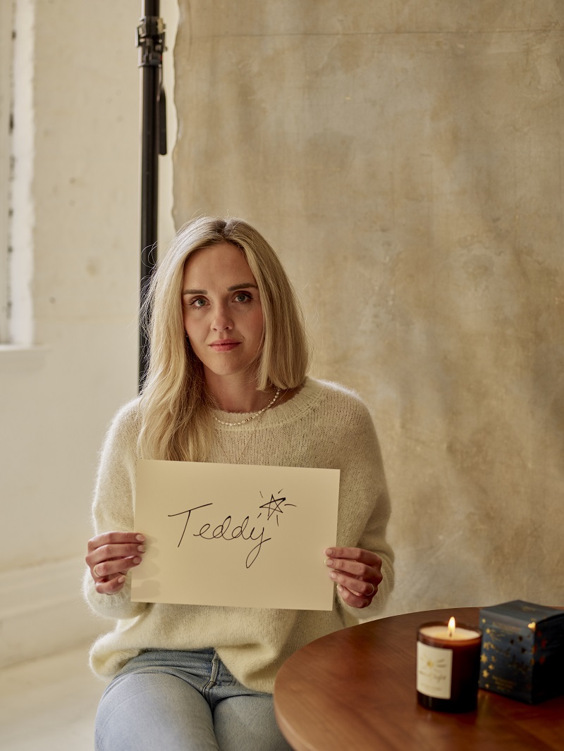 Author Elle Wright wears a cream coloured jumper and jeans. She holds up a sign with the name of her son Teddy on it, she's sitting next to a lit candle