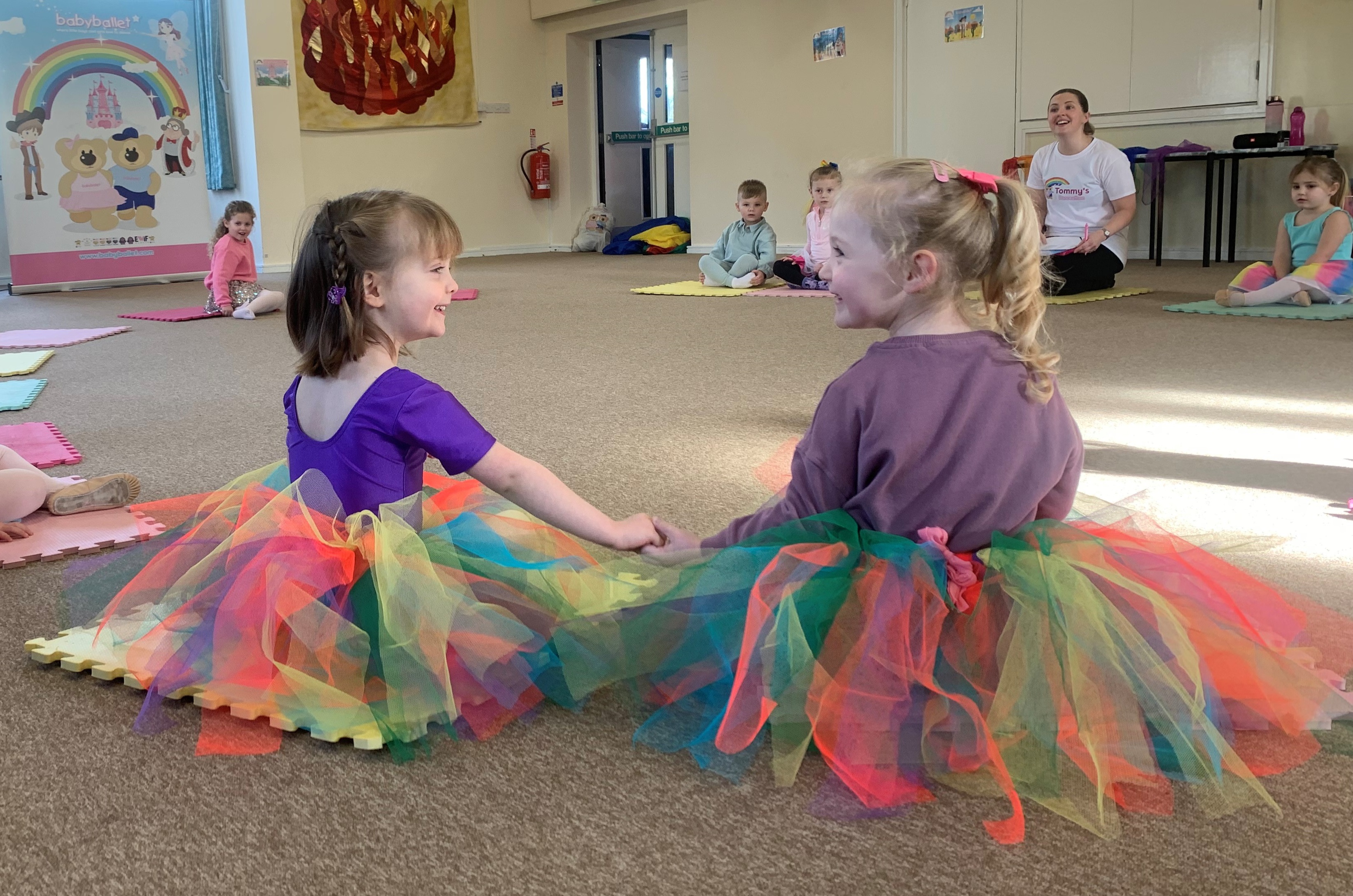 Two little girls in big rainbow tutus sit on the floor facing toward each other smiling