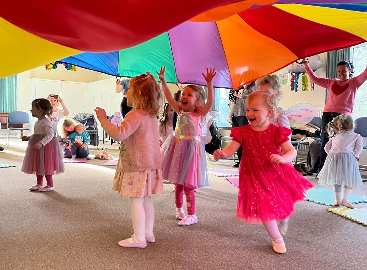 Little dancers happily run around under a big rainbow sheet held up by the grown-ups
