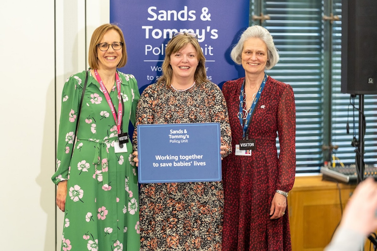 Three women standing in front of a Sands & Tommy's banner holding a sign committing to saving babies' lives