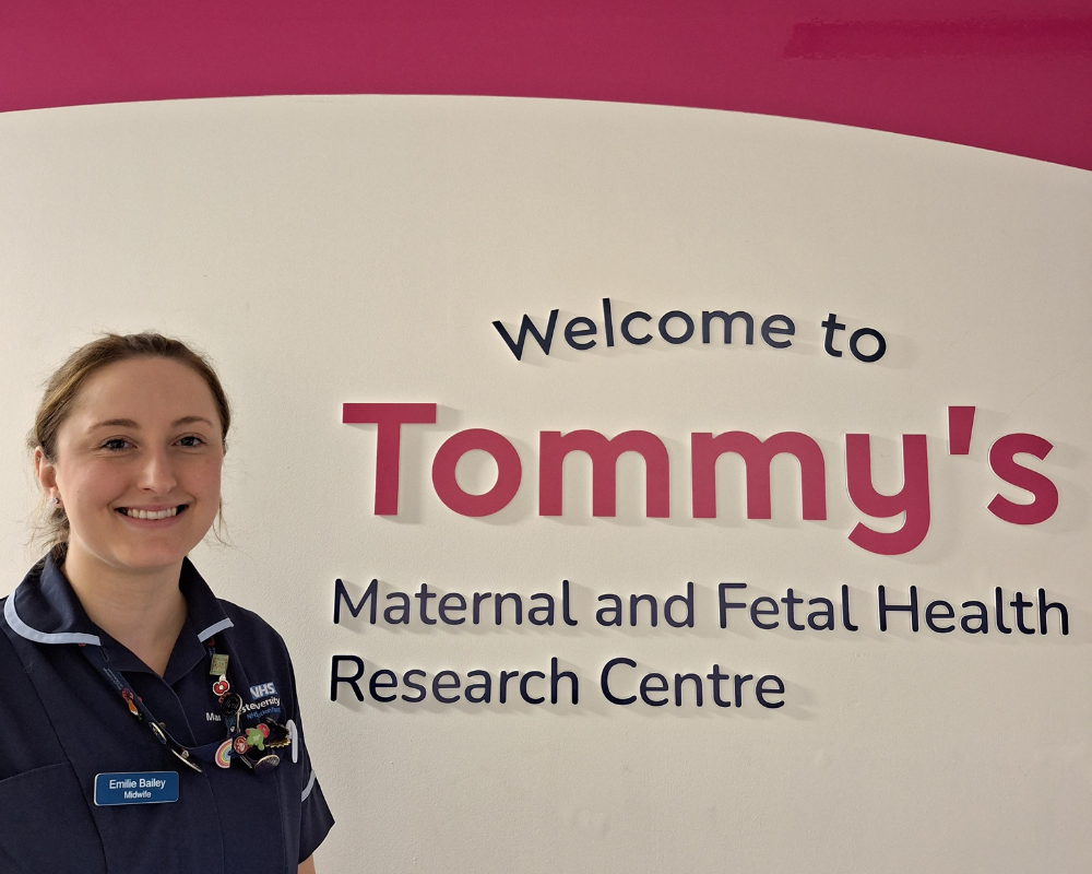 Emilie stood smiling next to a sign that reads: Welcome to Tommy's Maternal and Fetal Health Research Centre