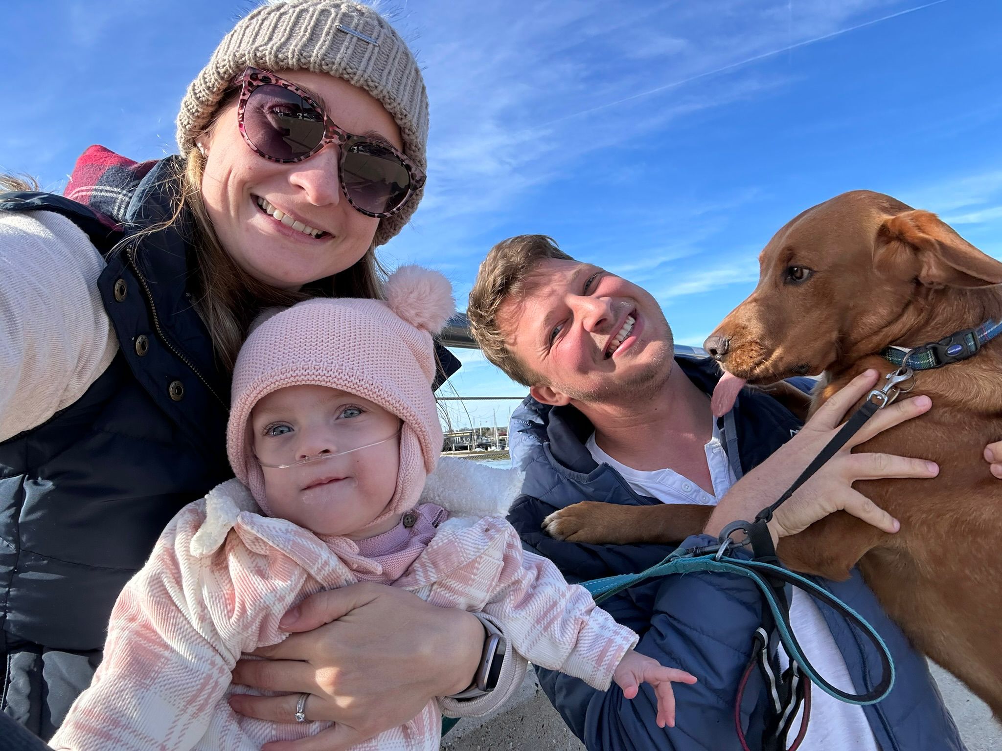 A family - mother, father, young daughter and their dog - smile at the camera outside against a blue sky.
