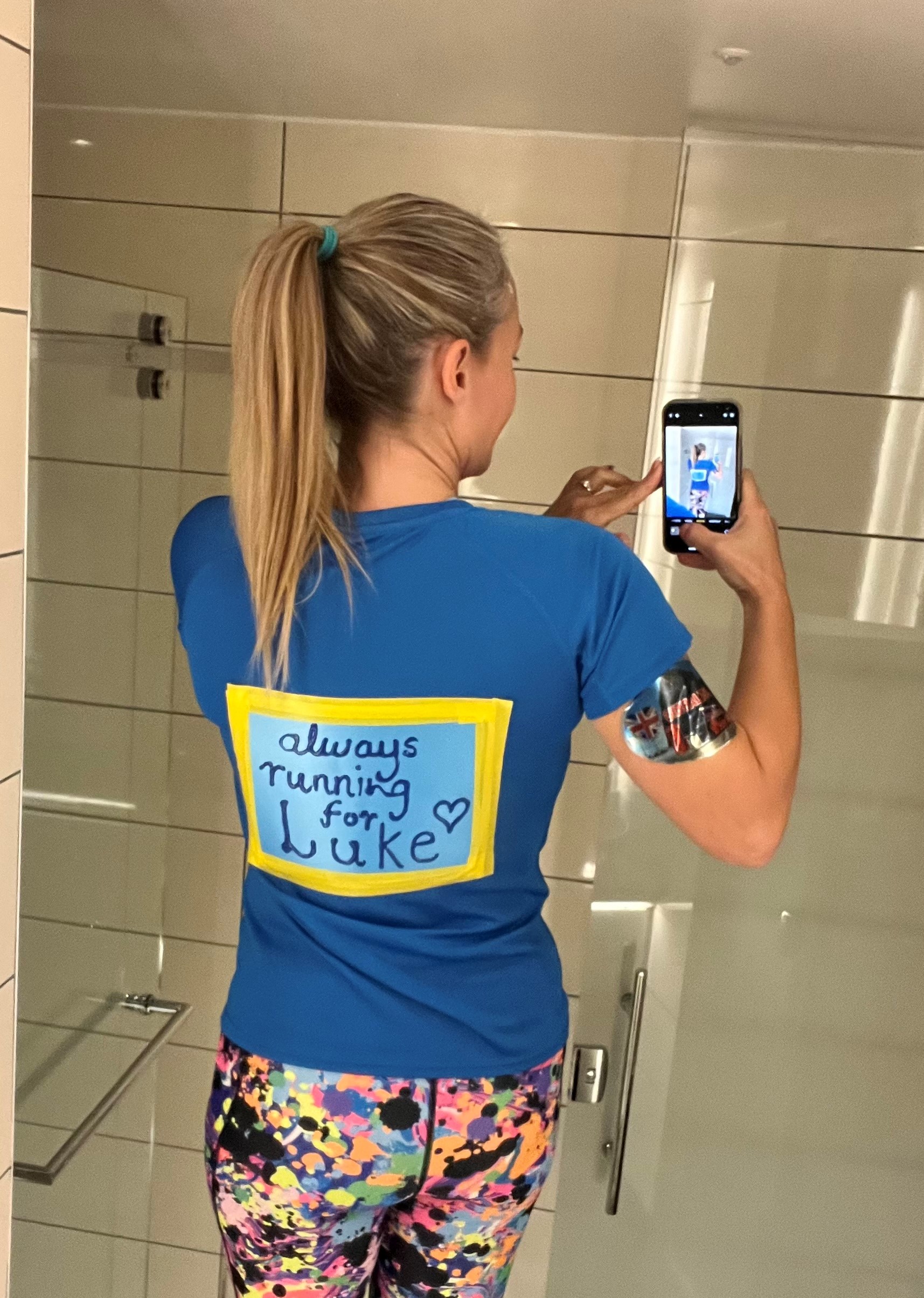 A woman in running clothes is taking a mirror selfie over her shoulder to show the back of her t-shirt, which reads 'Always running for Luke'.